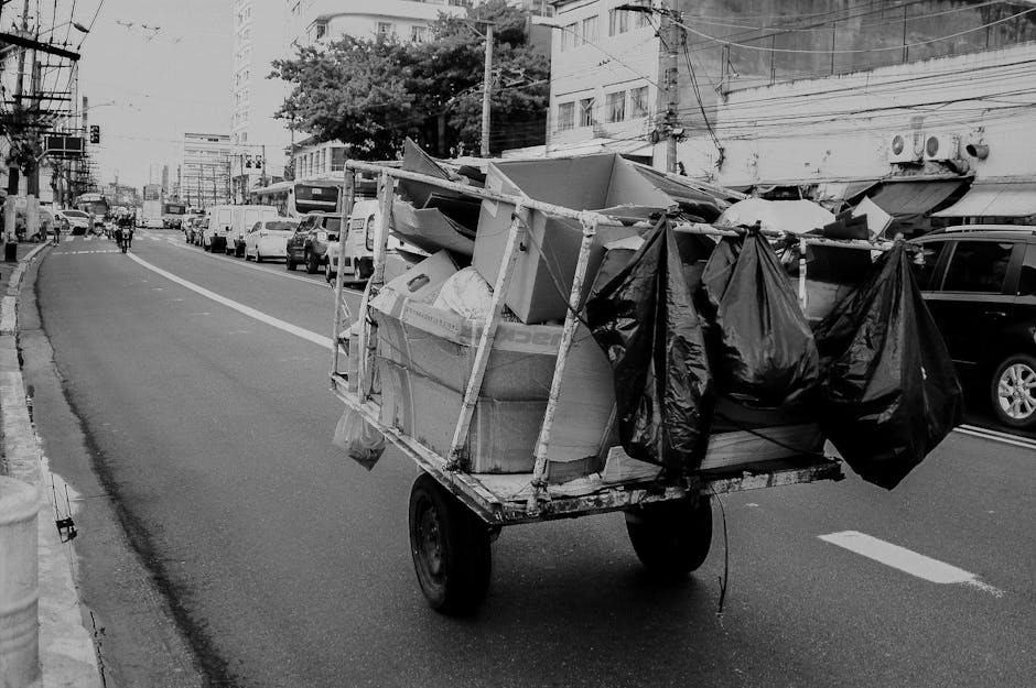 A small, homemade mobile cart with two black plastic bags hanging from its sides is seen on a city street, loaded with various discarded items including large cardboard boxes, a piece of furniture, and other household debris. The cart appears to be constructed from wooden planks and metal, with a single axle and two wheels, positioned on the asphalt roadway close to the curb. Surrounding the scene are parked cars, multi-storey buildings, and overhead power lines, indicating an urban environment. The street features a gentle curve, with a few pedestrians and vehicles visible in the background, suggesting recent or ongoing rubbish collection activity possibly linked to private waste handling or independent clearance services. This scene captures a typical instance of on-site waste accumulation, potentially part of a local refuse removal or rubbish disposal process managed outside municipal collection routines. The environment is well-lit with natural daylight, emphasizing the contrast of the waste and cart against the background of city life, aligning with professional rubbish removal service contexts.