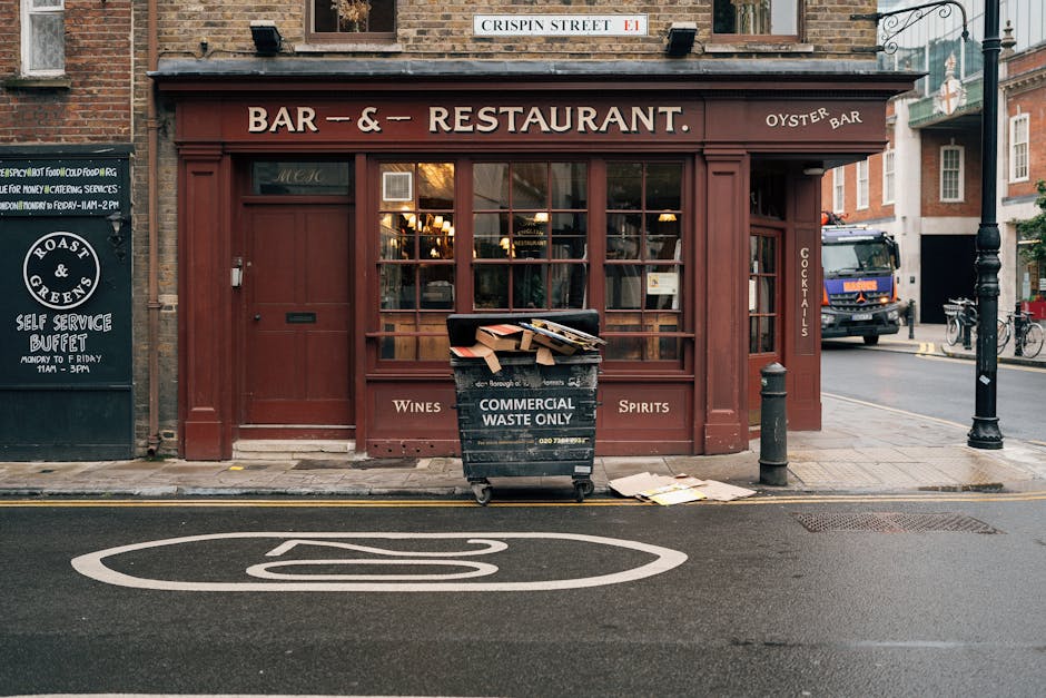 A black wheeled bin labeled 'Commercial Waste Only' positioned on the sidewalk in front of a red-brick building housing a bar and restaurant. The bin is filled with flattened cardboard boxes and paper waste, some of which are protruding from the top. The concrete pavement surrounding the bin has a few loose sheets of paper nearby. To the left, a black sign advertises a self-service buffet and food offerings, while to the right, a metal bollard prevents vehicle access to the pavement. A small metal post with a chain is attached to a nearby streetlamp pole, which is situated on the corner of a street with a painted bus stop lane on the asphalt. Behind the restaurant, part of a wide street is visible, including parked bicycles, a truck, and neighboring buildings. The scene appears to be during daylight hours with natural ambient lighting, typical of an urban environment where independent waste collection services like those offered by Rubbish Removal Notting Hill might handle rubbish clearance for local businesses such as this one.