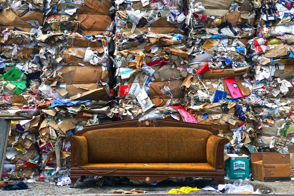 In the foreground, an antique-style sofa with a wooden frame and patterned fabric upholstery in shades of brown and gold is positioned on a paved surface. Behind it, a large, uneven pile of mixed waste dominates the scene, consisting of cardboard boxes, crumpled paper, plastic packaging, and other paper-based rubbish in various colors, including brown, white, red, blue, and green. The cardboard appears worn, some torn and flattened, while the paper and plastic waste are loosely stacked, creating a chaotic and cluttered backdrop. To the right of the sofa, a cardboard box labeled 'Sista' and other smaller containers are visible amongst the rubbish, indicating possible disposal or collection points. The environment is outdoors with diffuse daylight illuminating the scene, emphasizing the textures and colors of the waste and the vintage sofa. This image exemplifies a site where private rubbish disposal or waste collection services, such as those offered by Rubbish Removal Notting Hill, might be engaged to manage bulky waste accumulation, including furniture and mixed household rubbish, in an urban setting.