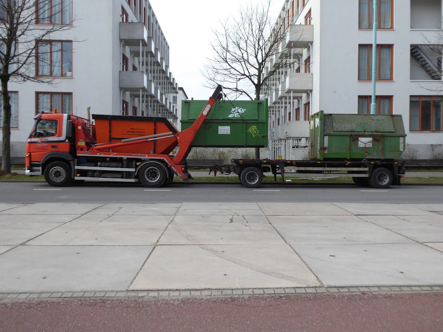 A large red rubbish disposal truck with an attached flatbed trailer is parked on a paved street in front of a multi-storey residential building. The truck features a hydraulic lifting mechanism positioned at the rear, which is engaged with two green metal skips or containers loaded onto the trailer. One container on the left is larger, with graffiti markings of a white bicycle symbol on its side, and appears to be partially filled with waste. The other smaller container on the right is also green, showing signs of wear and dirt, and has a closed lid. The scene is well-lit, with leafless trees on either side of the truck, indicating an urban environment during colder months. The surroundings include modern building facades with balconies and windows, and a street lamp pole near the right side of the image. The scene depicts an instance of private rubbish collection, indicative of alternative waste handling services like those provided by Rubbish Removal Notting Hill for efficient rubbish and furniture disposal.