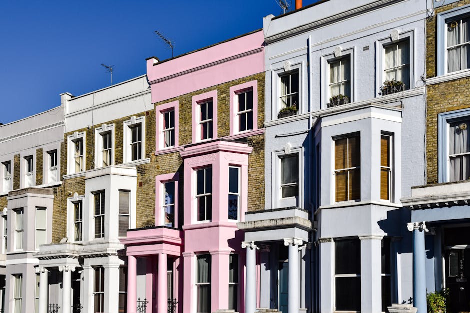 A row of Victorian terraced houses in Notting Hill, featuring bay windows, decorative moldings, and colorful facades in shades of blue, pink, and white. The buildings have brick exteriors with painted surfaces and are situated along a paved street. Some windows are adorned with flower boxes, and metal railings line the front steps. The image is taken during daylight hours, with natural light illuminating the façades and casting soft shadows on the street. The overall scene depicts typical residential architecture in an urban area, with no visible rubbish or waste collection activity. The orderly appearance and intact condition of the buildings suggest they are well-maintained and part of a thriving neighborhood, potentially serviced by private waste removal companies such as Rubbish Removal Notting Hill, specializing in alternative waste handling solutions like private rubbish collection or on-site clearance for residential properties.