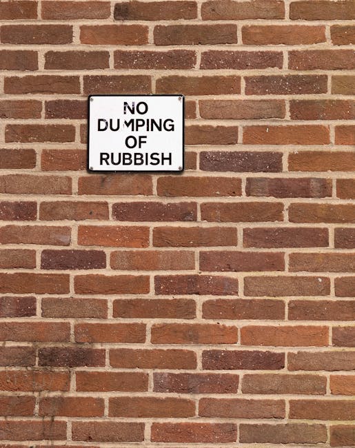 A close-up of a rectangular black and white metal sign mounted on a red brick wall. The sign displays the message 'NO DUMPING OF RUBBISH' in uppercase letters, with the word 'DUMPING' partially obscured or damaged. The brick wall behind the sign has a rough texture with reddish-brown and dark brown bricks arranged in a running bond pattern, and light-colored mortar joints. The sign is positioned in the upper left section of the image, with the wall extending below and to the right, creating a neutral, urban setting that relates to waste management and private rubbish collection services as offered by Rubbish Removal Notting Hill.