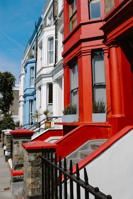 The image displays a row of Victorian-style terraced houses with distinct color schemes, featuring the front facades and staircases leading up to the entrances. The house on the right is painted in a bold, red warm hue with detailed architectural elements such as arched window frames, decorative columns, and a small staircase with a red handrail and concrete steps. The neighboring house on the left is painted in a softer, light blue shade with similar Victorian architectural features, including bay windows and ornate moldings. Both properties have small front gardens with potted plants positioned on the window sills and near the entrance. The staircases are constructed from concrete, with some steps showing signs of wear. In front of the houses, there is a low brick wall topped with a black metal fence, separating the properties from the pavement. The surroundings are well-lit by natural daylight, with a clear blue sky overhead, providing a bright and clear environment. Rubbish removal services, such as those offered by Rubbish Removal Notting Hill, may support these residents with on-site clearance or private waste collection, especially considering the urban setting and limited space for waste storage.