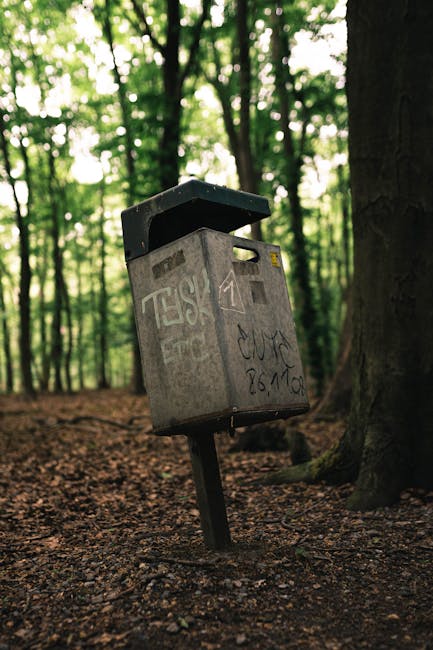 A weathered grey metal mailbox with a slightly tilted position stands on a dark metal post in a forested area, surrounded by tall trees with green leaves. The mailbox has a rectangular shape with a black, slightly curved hinged lid on top, and its surface is covered in graffiti and faded markings, giving it an aged appearance. The ground beneath is composed of compacted dirt and brown fallen leaves, contrasting with the lush, dense canopy in the background where sunlight filters through the tree branches, creating a dappled light effect. The scene suggests an outdoor setting away from urban environments, possibly near walking trails or nature paths. The presence of the mailbox amid these natural surroundings hints at informal or unconventional waste collection points, aligning with alternative rubbish handling practices sometimes employed in private or remote locations, supported by services like Rubbish Removal Notting Hill that handle waste outside conventional municipal routes.