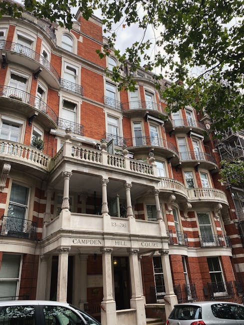 The image depicts a multi-storey residential building with a historic architectural style, featuring a central entrance supported by four classical columns and a balustrade balcony above. The building's façade is composed of red brickwork with contrasting white stone accents around the windows and decorative elements, creating a striking appearance. Several large windows with white frames are visible, some with small wrought iron railings, and the building extends upwards with multiple floors, each with bay windows and curved balconies. Green leaves from overhanging trees partially obscure the top of the structure, adding a natural element to the scene. The environment appears to be a city street with parked cars in the foreground, and the lighting suggests an overcast day with diffuse natural light. This scene exemplifies a typical urban residential property that could be subject to on-site clearance or private rubbish removal services, such as those offered by Rubbish Removal Notting Hill, to facilitate cleaning or maintenance of such historic buildings while supporting quality waste disposal alternatives in London.