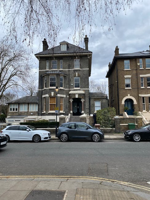 A tall, three-storey Victorian-style residential building with a dark brick exterior, white window frames, and decorative stone details around the bay windows and entrance. The front door is dark, accessed by a short flight of stairs with black railings, and there's a small, pitched roof above the main entrance. To the left, a lower extension with large windows and a pitched roof is visible. The building is situated on a city street with a row of parked cars, including a white convertible, a dark hatchback, and another dark vehicle, all parked alongside the pavement. In the background, leafless trees and a cloudy sky create an overcast atmosphere. The setting suggests an urban environment, typical of London residential districts, where private waste collection or rubbish removal services from companies like Rubbish Removal Notting Hill might be employed for clearing household waste following property clearance or renovation activities.
