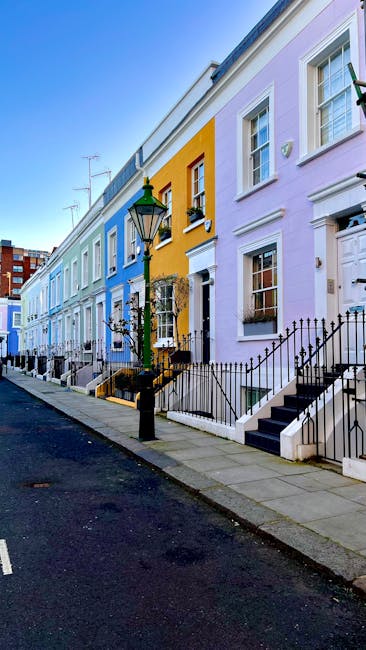 A row of colorful terraced houses lining a city street, each house painted in pastel shades including blue, yellow, and light purple. The properties feature white-framed sash windows, some with flower boxes beneath, and small front gardens enclosed by black iron railings. In the foreground, a black lamppost with a green lantern stands adjacent to a paved sidewalk. The steps leading up to the front doors are made of dark stone, contrasting with the light-colored facades. The street surface is dark asphalt with a white line marking the edge, and an electric pole with wires extends into the distance. The scene is illuminated by natural daylight, casting soft shadows and highlighting the vibrant, well-maintained exterior of the houses. The image suggests an urban residential area where private waste collection or on-site rubbish disposal services, such as those offered by Rubbish Removal Notting Hill, might be utilized in place of municipal rubbish collection, given the absence of visible large waste containers or communal skips.
