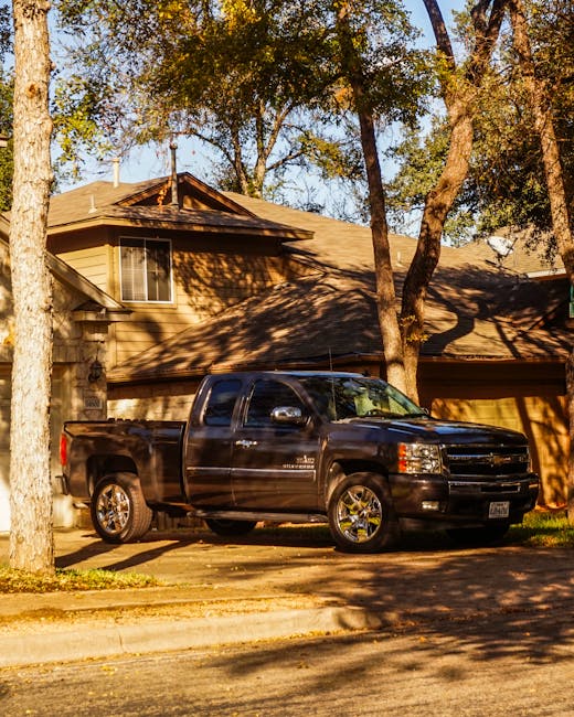 A black pickup truck parked in front of a residential house with beige siding and a dark brown roof. The truck, situated on a paved driveway, has a glossy finish and chrome wheels that reflect sunlight. The house features a large window with white trim on the upper floor, and the surrounding area is shaded by tall trees with green and some golden leaves, indicating an autumn setting. Shadows from the trees fall across the driveway and part of the truck. The environment appears quiet and suburban, with a natural light atmosphere suggesting late afternoon. This scene illustrates an example of private waste handling or preparation for rubbish removal services, such as those provided by Rubbish Removal Notting Hill, which often involves on-site clearance like this for efficient rubbish collection or disposal.