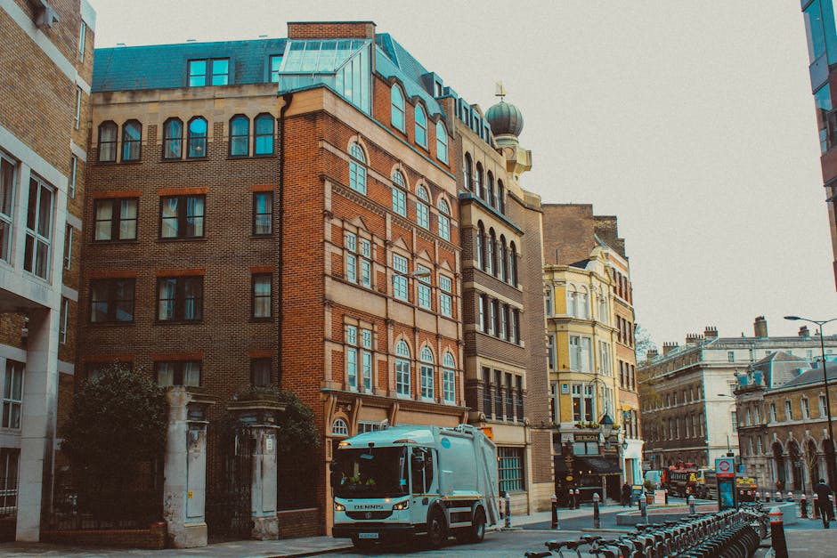 A city street scene featuring a mix of historic and modern buildings, primarily constructed from red and yellow brick with large windows and decorative architectural details. In the foreground, a blue waste collection truck is parked on the pavement, slightly to the left of the center, with its side facing the viewer. The truck appears to be a commercial vehicle used for private rubbish collection, in front of a gated entrance flanked by stone columns and greenery. Surrounding the truck are several metal bike racks and bollards that segment the pedestrian area from the road. The background features additional buildings with varied facades, including one with a rounded, domed turret, and another with classic detailing and ornate features. The sky is overcast, casting diffuse natural light over the scene, emphasizing the textures of the brickwork and the metallic sheen of the waste collection vehicle. This image illustrates an urban environment where independent rubbish removal services operate alongside city infrastructure, highlighting the relevance of private waste handling methods in densely built areas near residential and commercial properties, consistent with services offered by Rubbish Removal Notting Hill.