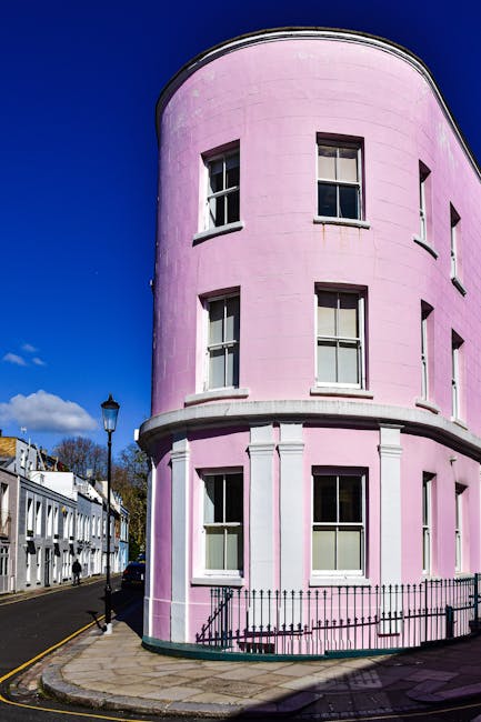The image displays a three-story, rounded corner building painted in pastel pink with a smooth, curved facade. The building features white window frames with multiple-pane sash windows, each with dark glass panes reflecting the sky. The ground floor has larger windows, and the upper floors have smaller, evenly spaced windows aligned vertically. Surrounding the building is a narrow sidewalk paved with flat stone slabs, bordered by a black metal fence that encloses a small outdoor area at the front. To the left, part of a street with parked cars is visible, along with a traditional black lamppost near the corner of the building. In the background, there are more white terraced houses with dark roofs, and a bright blue sky with a few scattered clouds. The scene is illuminated by natural daylight, with shadows cast on the building's surface. This architectural setting, with its clean and orderly appearance, complements the context of professional waste and rubbish removal services, such as those offered by Rubbish Removal Notting Hill, which often handle clutter associated with residential and commercial properties in urban areas.
