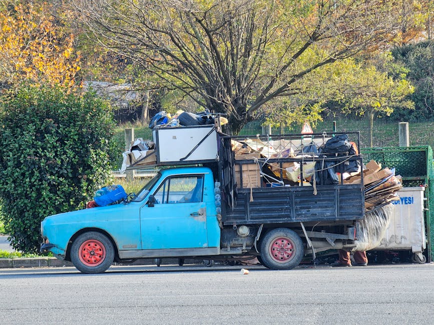 A small old blue flatbed truck parked on the side of a paved road, loaded with a large pile of mixed waste materials including flattened cardboard boxes, black plastic garbage bags, and various miscellaneous debris. The truck's body is visibly weathered with some paint chipping, and it has red-painted wheels. Behind the truck, there is a dense green hedge with a large tree featuring sparse, yellow-green leaves, indicating an autumn setting. The surrounding environment appears to be a suburban or residential area, with a metal fence partially visible in the background. The overall scene suggests a private waste collection or rubbish removal activity, possibly managed by Rubbish Removal Notting Hill, and illustrates an on-site clearance of bulky waste or household rubbish outside a property.