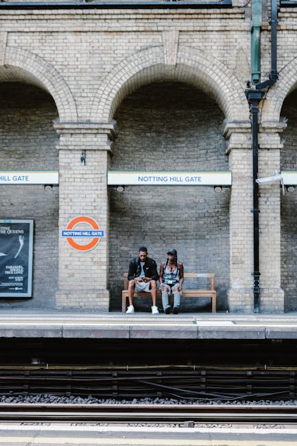A brick platform at Notting Hill Gate Underground Station features two young individuals sitting on a wooden bench, with the male on the left dressed in a black jacket, grey shorts, and white sneakers, and the female on the right wearing a black and white patterned top, grey pants, and black sneakers. The background displays the station's brick wall structure with two large arches, and signs indicating 'NOTTING HILL GATE' in bold blue lettering. The station sign with the roundels and the station name is positioned centrally below the arches, and a black drainage pipe runs vertically along the right side of the wall. The platform surface appears to be made of light grey concrete, and the edge of the platform is visible along the bottom of the image, with railway tracks in the foreground. The lighting suggests natural daylight, with shadows cast by the station's structure. The scene is calm and depicts an everyday moment at this transportation hub, illustrating the typical environment for passengers waiting for a train. This setting can relate to private or alternative waste handling experiences, such as when passengers discard rubbish or await discharge in public spaces, making it relevant to rubbish removal services like those provided by Rubbish Removal Notting Hill.