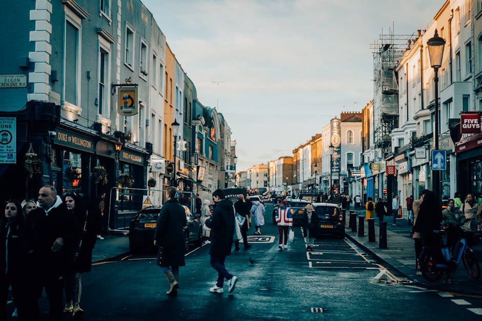 A busy urban street scene during late afternoon, featuring a row of multi-storey shops with varying facades, including a pub called 'Duke of Wellington' on the left. The buildings display a mix of architectural styles, some with scaffolding indicating ongoing maintenance. Pedestrians, dressed in casual and business attire, walk along the pavement, while others are gathered near the shop entrances or waiting to cross the street. Several cars and taxis are parked along the curb, with a black vehicle in the center of the street. Street furniture includes bollards, lampposts, and road signage, including a blue one-way arrow sign on the right. The pavement appears wet, possibly from recent rain, reflecting the warm light of the setting sun. The overall atmosphere is lively yet orderly, typical of a vibrant commercial area, reflecting the context of alternative waste collection and urban rubbish management through frequent pedestrian and vehicular activity in a typical city environment. Rubbish removal companies like Rubbish Removal Notting Hill might operate in this context to manage waste from such busy streets effectively, particularly with on-site clearance or private disposal services.