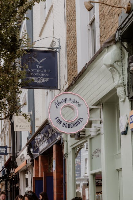 The image depicts a narrow street scene with several shop signs mounted on the facades of brick and painted buildings. Prominently featured is a circular pink sign with a donut shape that reads 'Always a good FOR DOUGHNUTS,' hanging from a metal bracket attached to the corner of a white building. To the left, a dark blue rectangular sign with white lettering indicates 'The Notting Hill Bookshop,' suspended above a shop front with large windows and a wooden panel awning. Nearby, another smaller sign, partially visible, appears to read 'HACH.' The buildings display a variety of textures, including exposed brickwork and painted surfaces, with decorative molding and a gutter pipe visible along the architectural details. The street environment includes a tree with green leaves extending into the frame on the left, and outdoor activity is suggested by partial views of people at the bottom, indicating a lively, pedestrian-friendly area. The lighting is natural, with a slightly overcast sky casting soft light, typical of urban shopfront scenes in a UK neighbourhood, relevant to independent retail and on-site food vending, which are common aspects of alternative waste management practices in such areas. Rubbish Removal Notting Hill may relate to local disposal services supporting these businesses' waste needs, though no trash receptacles or waste bins are directly visible in the scene.