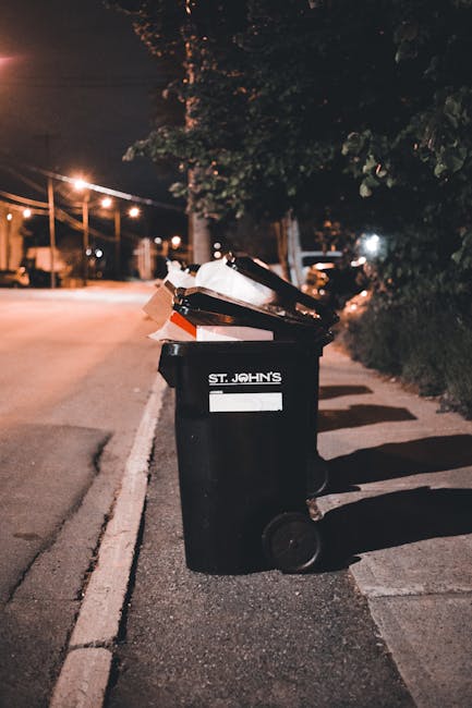 A black plastic wheelie bin labeled 'St. John's' is positioned on the sidewalk beside a kerb on a quiet urban street at night. The bin is filled with various types of waste including cardboard boxes, plastic bags, and other rubbish, some of which are leaning or protruding from the open lid. The bin appears to be a typical waste collection container used for household or commercial rubbish. The street is illuminated by nearby streetlights that cast a warm glow on the scene, creating soft shadows on the pavement and the bin. In the background, there are trees and foliage, as well as faint outlines of parked cars and buildings, indicating a residential or mixed-use area. The environment suggests a setting for private waste disposal, possibly in preparation for an independent rubbish collection service, reflecting the context of alternative waste handling options outside of local authority collection schedules.